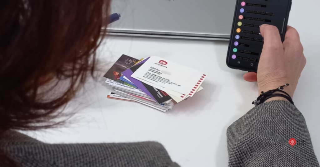 Una persona sentada frente a una mesa blanca en la oficina sostiene un teléfono móvil con una lista de contactos en la pantalla. Sobre la mesa hay varias tarjetas de visita apiladas. La imagen muestra el momento posterior a una feria, cuando se revisan contactos y tarjetas.
